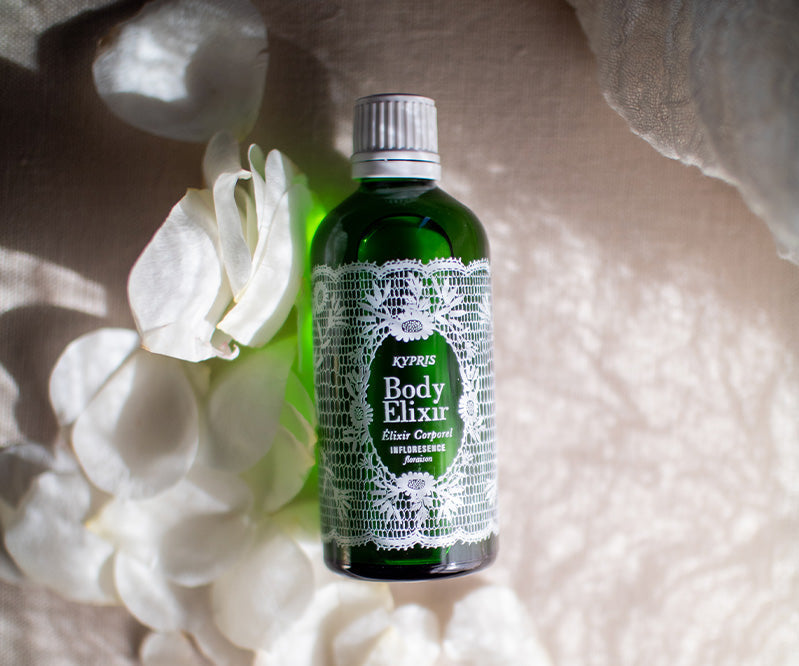 KYPRIS Body Elixir in green bottle with lace label, surrounded by white rose petals