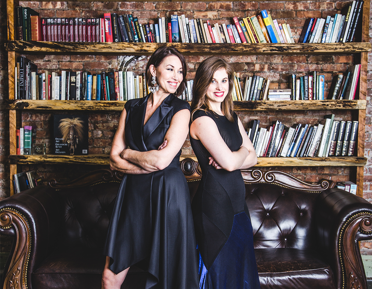 Two women in black dresses standing confidently in front of a bookshelf with leather couch