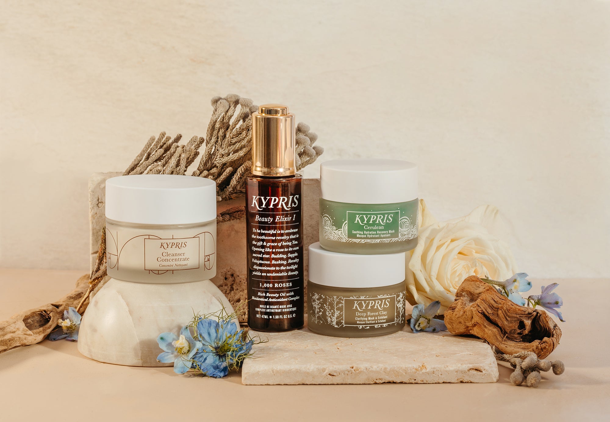 KYPRIS Cleanser Concentrate, Beauty Elixir I, Cerulean, and Deep Forest Clay skincare products with flowers and driftwood