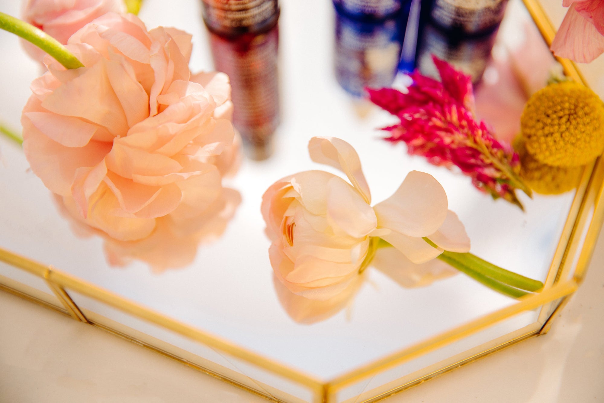 Pink peonies and colorful flowers on a gold-trimmed mirrored tray with skincare bottles in background