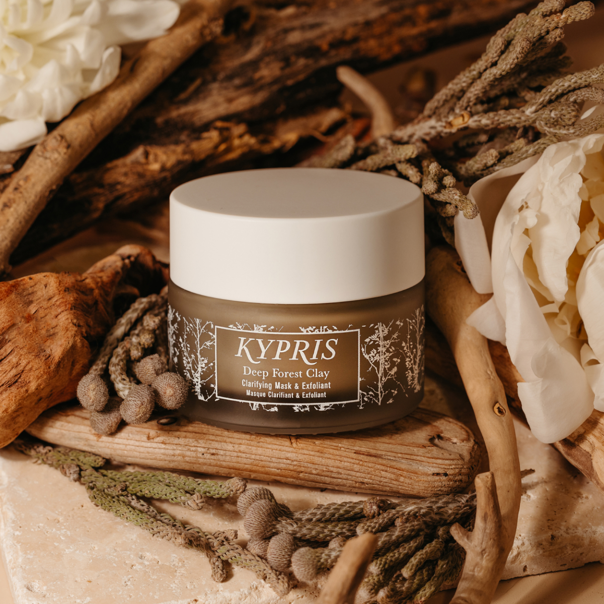 KYPRIS Deep Forest Clay Clarifying Mask & Exfoliant in earthy jar with white lid, surrounded by driftwood and white flowers