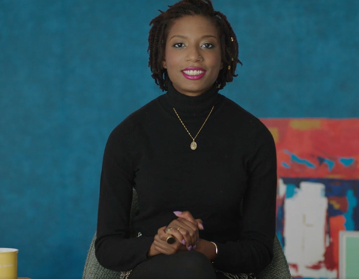 Woman in black turtleneck smiling, seated against teal wall with colorful abstract art, gold necklace, hands clasped