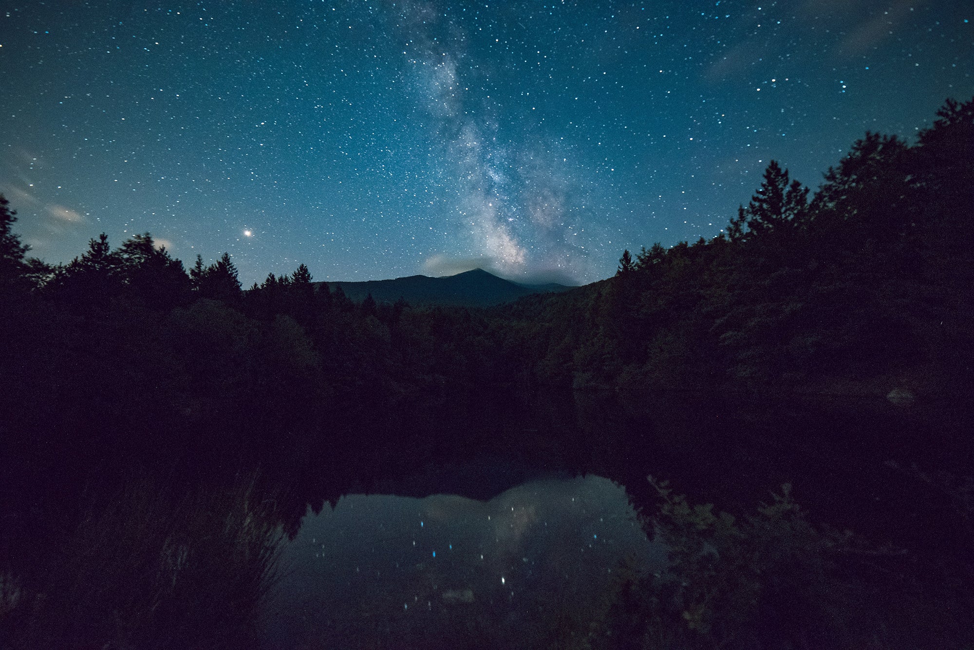 Starry night sky over forest with Milky Way reflected in calm lake